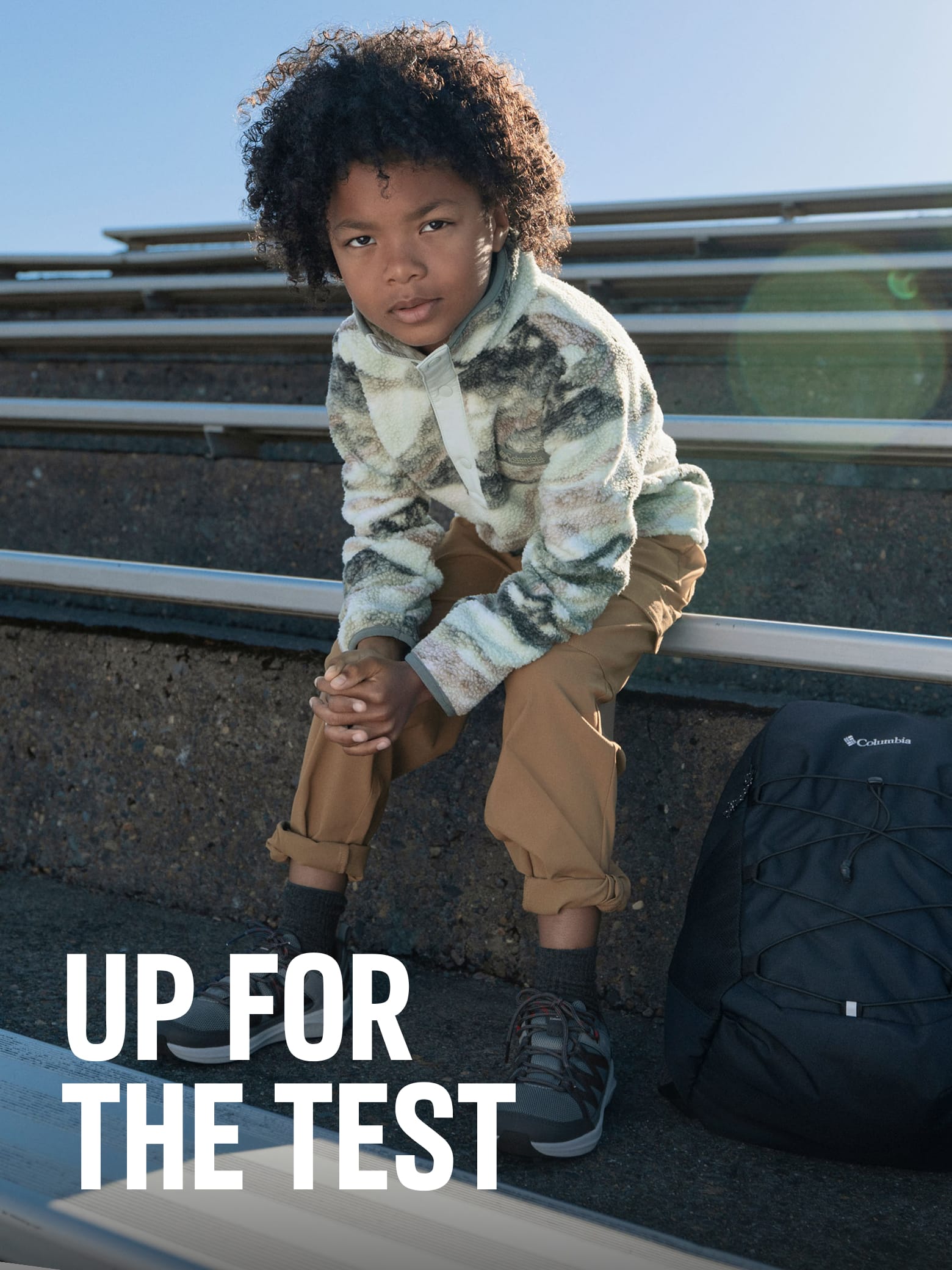 A little boy sitting on bleachers with text: UP FOR THE TEST