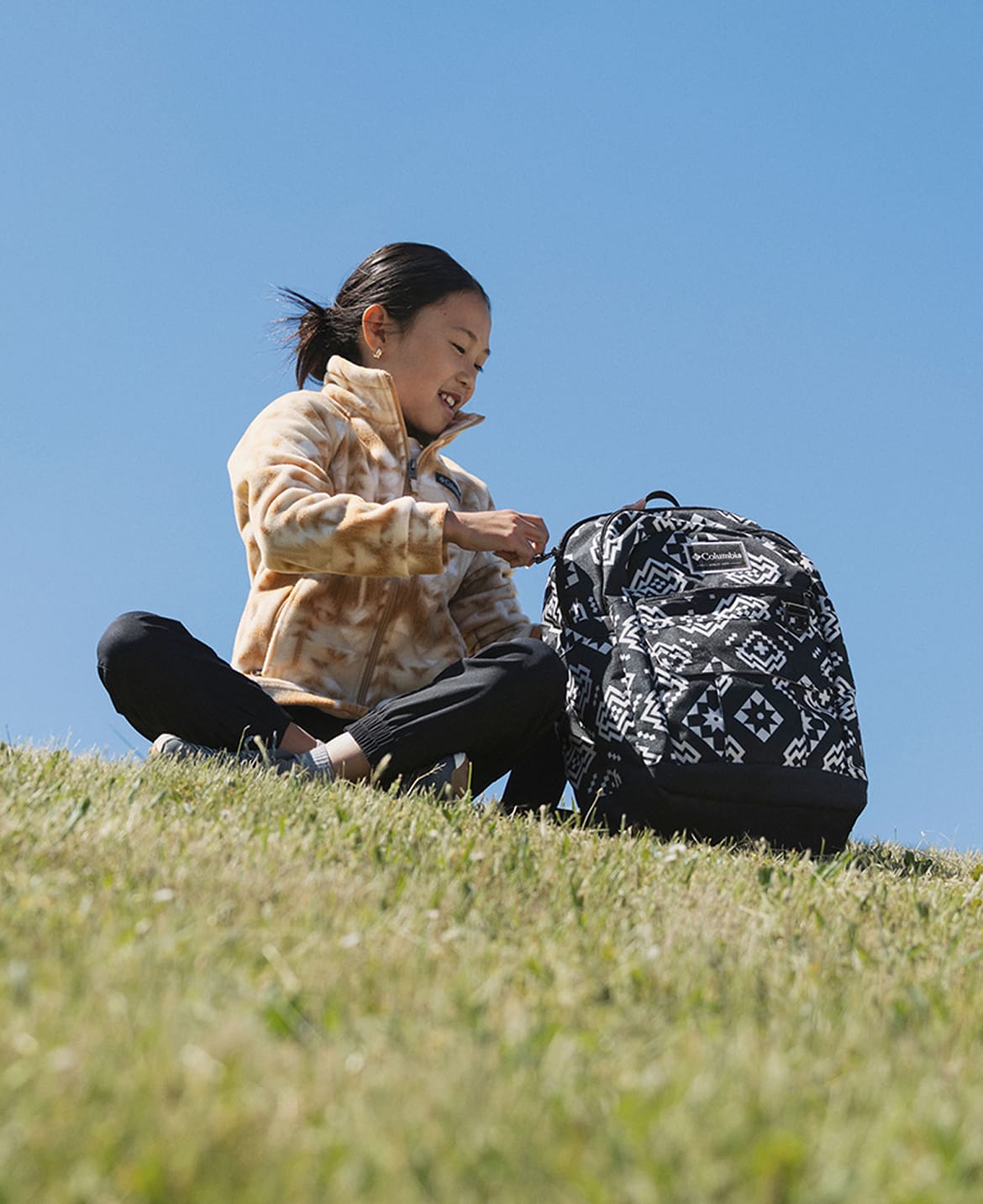 A little girl sitting on a grassy hill looking into her backpack.