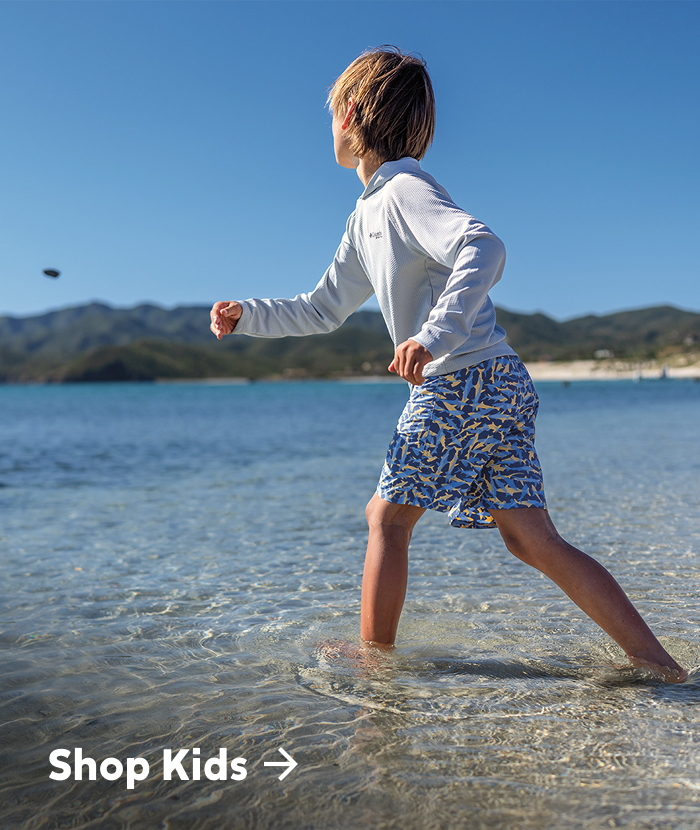 Shop Kids. A child throws a rock into shallow waters on a beach. 