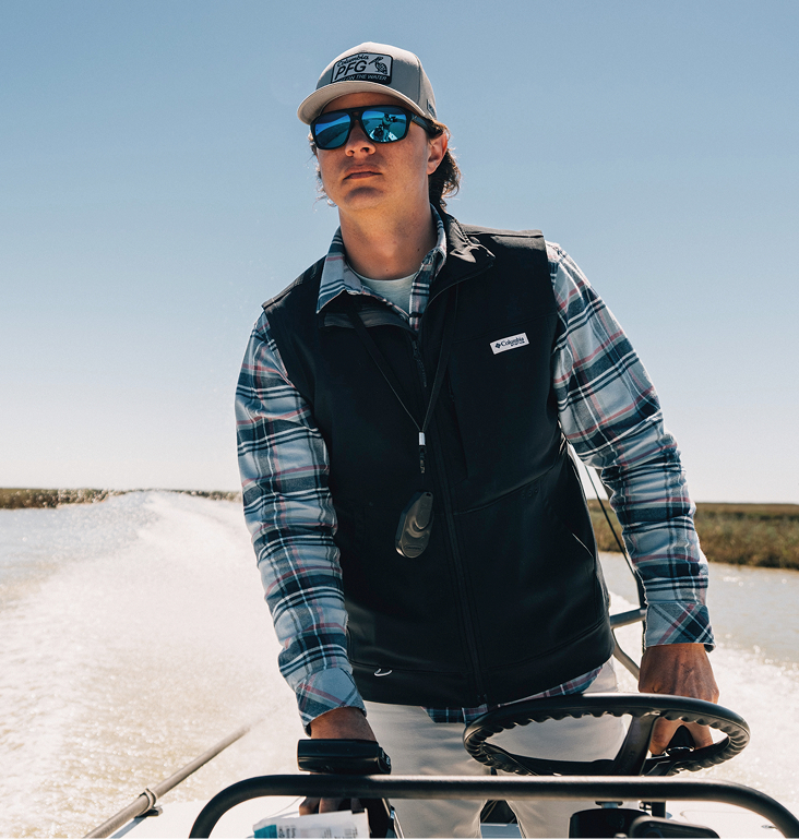 A man drives a boat while wearing a Columbia PFG vest over his flannel shirt. 